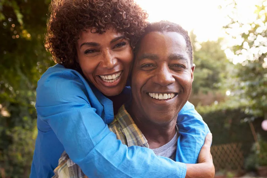 Autoimmune-Doctor A middle-aged black couple outside, the woman embracing the man from behind pressing her cheek to his, both are smiling due to successful Autoimmune Disorder treatment from Dr. Robert Powers of Eternal Vitality Orlando in Orlando.