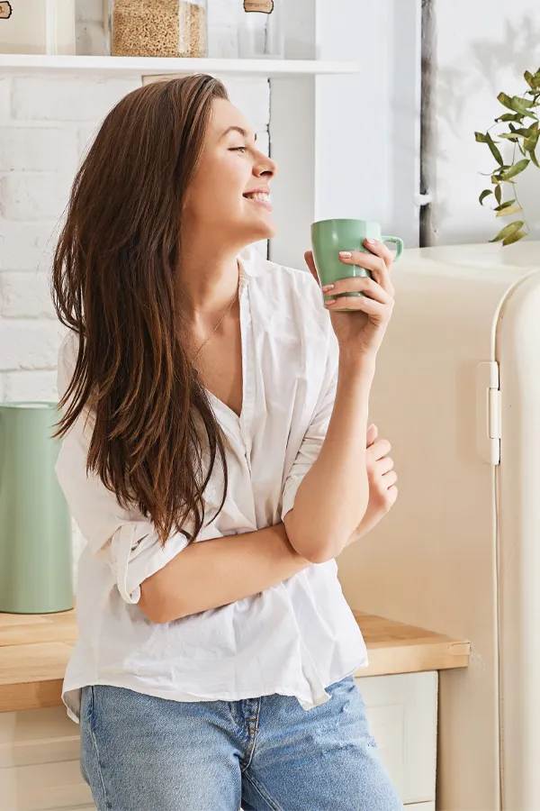 Crohns-Disease-Treatment A woman with a white blouse an jeans drinking tea in her kitchen, smiling after getting treatment for Crohn's Disease from Dr. Robert Powers of Eternal Vitality Orlando in Orlando.