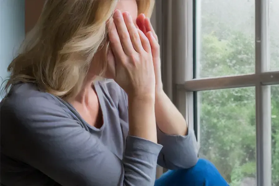 Graves-Disease-Clinic A blonde woman in a gray shirt sits in front of a window on a rainy day covering her eyes with her hands. Get treatment for Grave's Disease from Dr. Robert Powers of Eternal Vitality Orlando in Orlando.