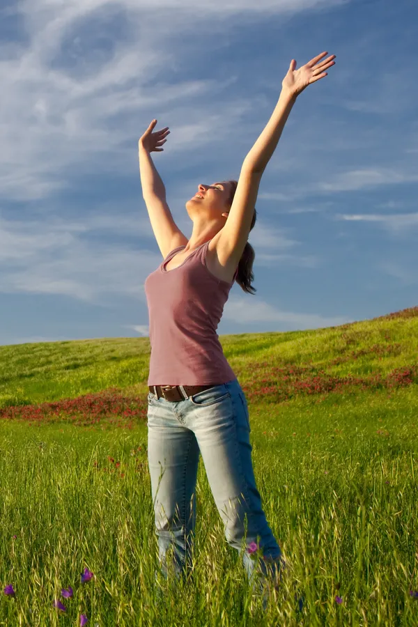 Graves-Disease-Treatment A woman in a tank top and jeans looks up with her arms outstretched to the sky in a field after successful treatment for Grave's Disease from Dr. Robert Powers of Eternal Vitality Orlando in Orlando.