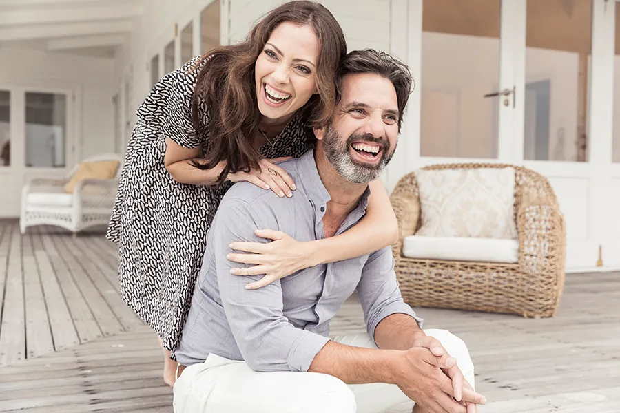 A middle-aged couple on a wooden porch with wicker furniture; the woman is playfully hugging the man from behind. They are benefiting from the health and wellness services from Dr. Robert Powers of Eternal Vitality Orlando in Orlando.