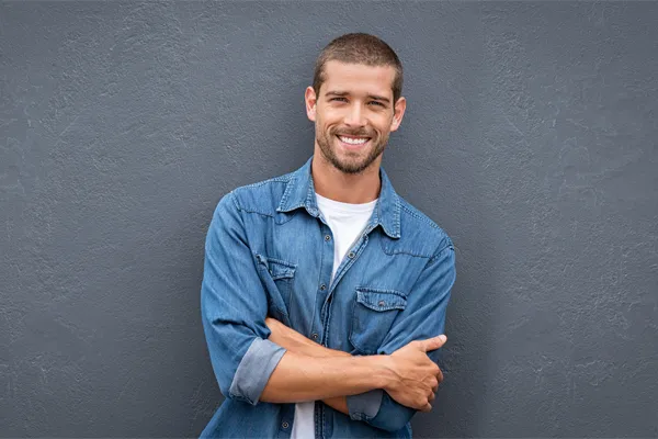 Hormones-for-Men-Doctor A man in a denim shirt stands smiling against a gray-blue wall, pleased with his testosterone hormone therapy from Dr. Robert Powers of Eternal Vitality Orlando in Orlando.