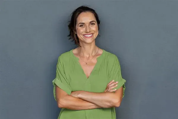 Hormones-for-Women-Doctor A woman in a green shirt stands smiling against a gray-blue wall, pleased with her hormone therapy from Dr. Robert Powers of Eternal Vitality Orlando in Orlando.