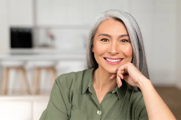 Menopause-Clinic An asian woman with long gray hair and a green shirt sitting and smiling on her couch, happy with her menopause care from [NAME} in Orlando.