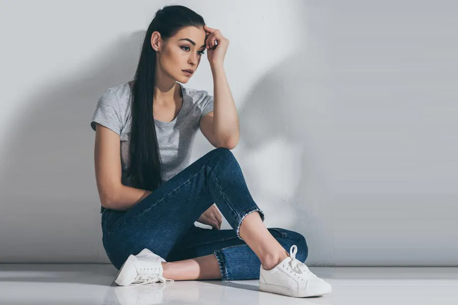 PCOS-Doctor A young brunette woman in a gray t-shirt and jeans sits against a gray wall with her head resting on her hand and a distressed expression. Get help for Polycystic Ovarian Syndrome (PCOS) from Dr. Robert Powers of Eternal Vitality Orlando in Orlando.