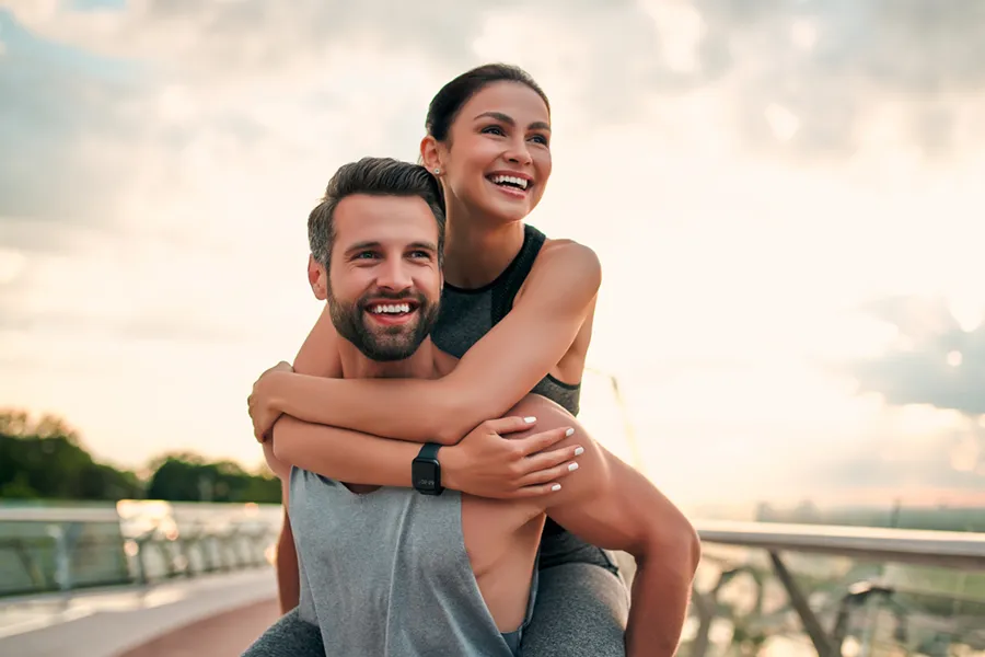 Radiofrequency-Body-Sculpting-Clinic A middle-aged couple in exercise clothes on a jogging trail; the woman is riding piggyback on the man playfully. Get Radiofrequency Body Sculpting treatments from Dr. Robert Powers of Eternal Vitality Orlando in Orlando.